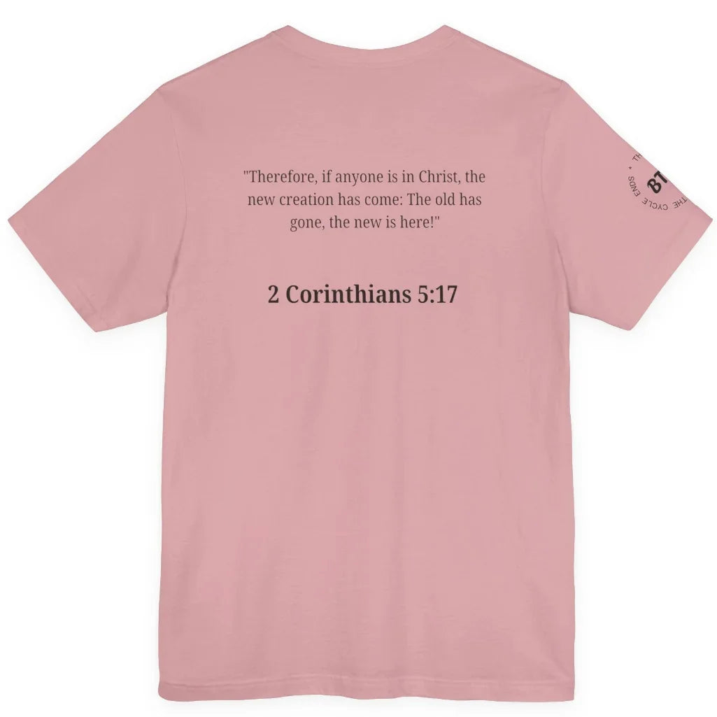 A orchid colored short-sleeve t-shirt shown from the back, featuring a black printed Bible verse from 2 Corinthians 5:17 in the center and a small circular graphic on the left sleeve.