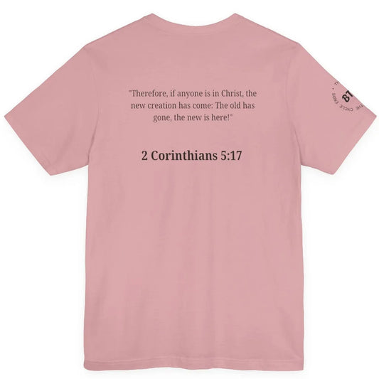 A orchid colored short-sleeve t-shirt shown from the back, featuring a black printed Bible verse from 2 Corinthians 5:17 in the center and a small circular graphic on the left sleeve.