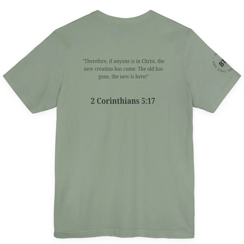 A sage green short-sleeve t-shirt shown from the back, featuring a black printed Bible verse from 2 Corinthians 5:17 and a small circular graphic on the left sleeve, displayed against a solid white background.