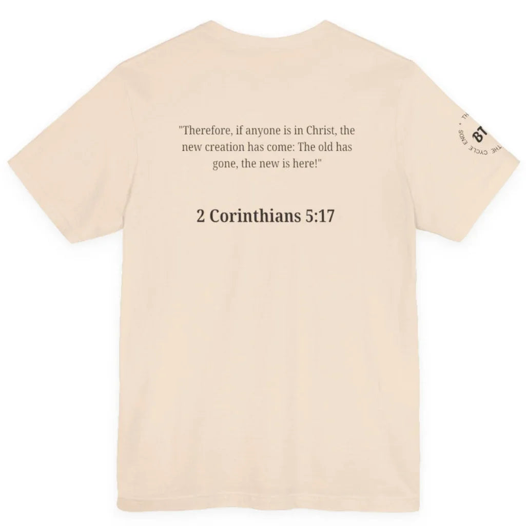 A light beige crewneck t-shirt shown from the back, featuring a black printed Bible verse from 2 Corinthians 5:17 in the center and a small circular graphic on the left sleeve.