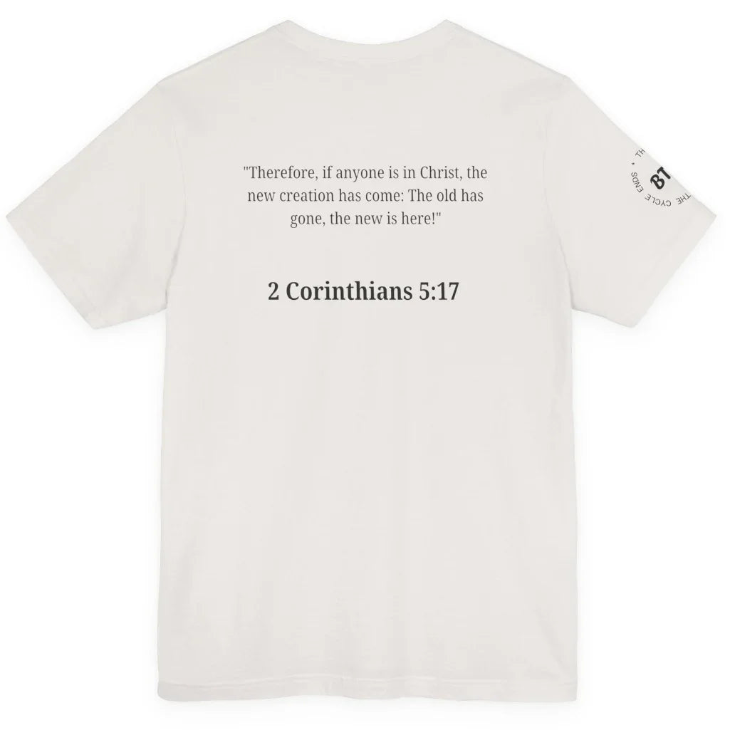 An off-white short-sleeve t-shirt shown from the back, featuring a black printed Bible verse from 2 Corinthians 5:17 in the center and a small circular graphic on the left sleeve.