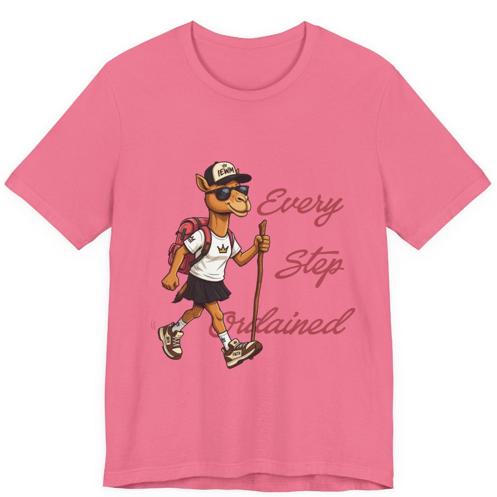 A pink short-sleeve crewneck t-shirt featuring a cartoon camel hiker graphic with the text 'Every Step Ordained' printed on the front, displayed against a solid white background.