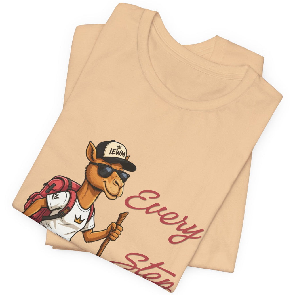 A folded light beige t-shirt with a cartoon camel graphic wearing a black cap that says 'IEWM' and sunglasses, holding a staff. The text 'Every Step' is printed in a dusty pink script on the right side of the shirt.
