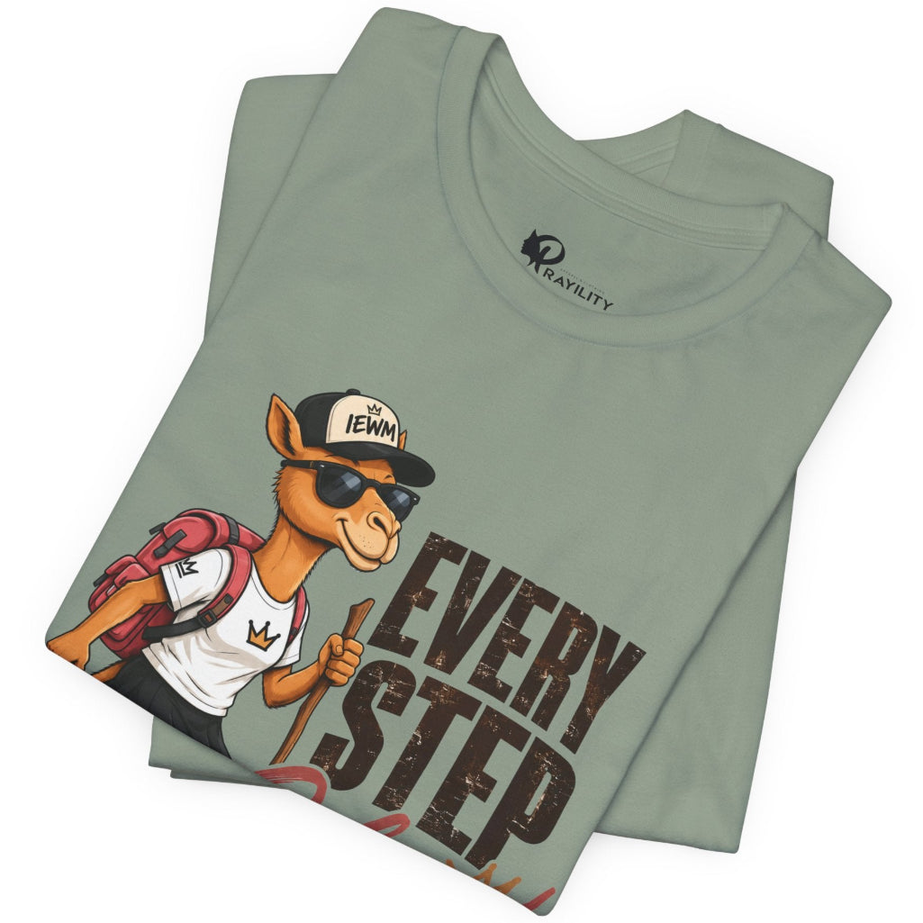 A folded sage green t-shirt featuring a cartoon camel hiker graphic with the text 'EVERY STEP' and a 'TEWNY' cap, displayed on a clean white background.