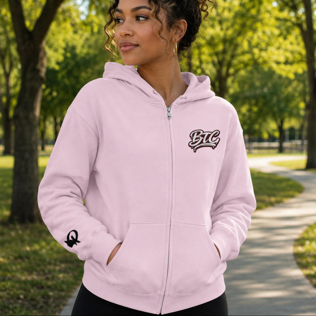 A woman wearing a light pink zip-up hoodie with a black and white 'BTC' graphic on the left chest and a small black graphic on the right sleeve, standing in a sunny park with trees and a pathway in the background.