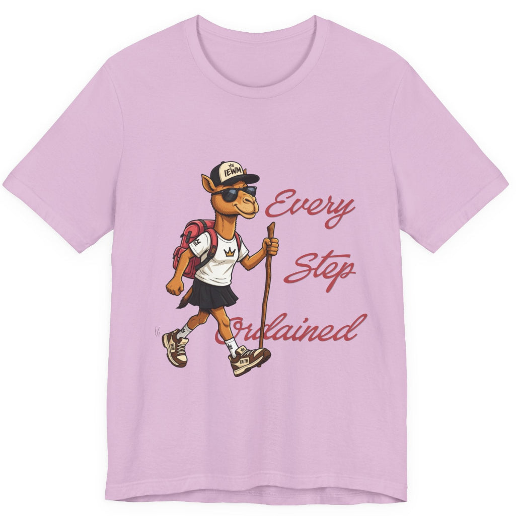A lilac short-sleeve t-shirt with a graphic of a cartoon camel hiker wearing a white shirt, black skirt, and a backpack, next to the cursive text 'Every Step Ordained'.