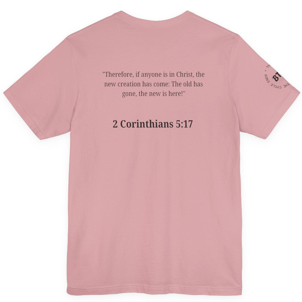 A orchid colored short-sleeve t-shirt shown from the back, featuring a black printed Bible verse from 2 Corinthians 5:17 in the center and a small circular graphic on the left sleeve.