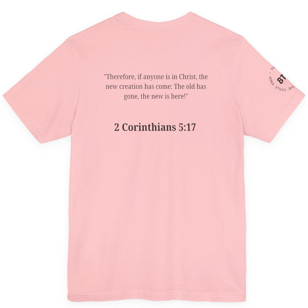 A light pink short-sleeve t-shirt shown from the back, featuring a black printed Bible verse from 2 Corinthians 5:17 in the center and a small circular graphic on the left sleeve.