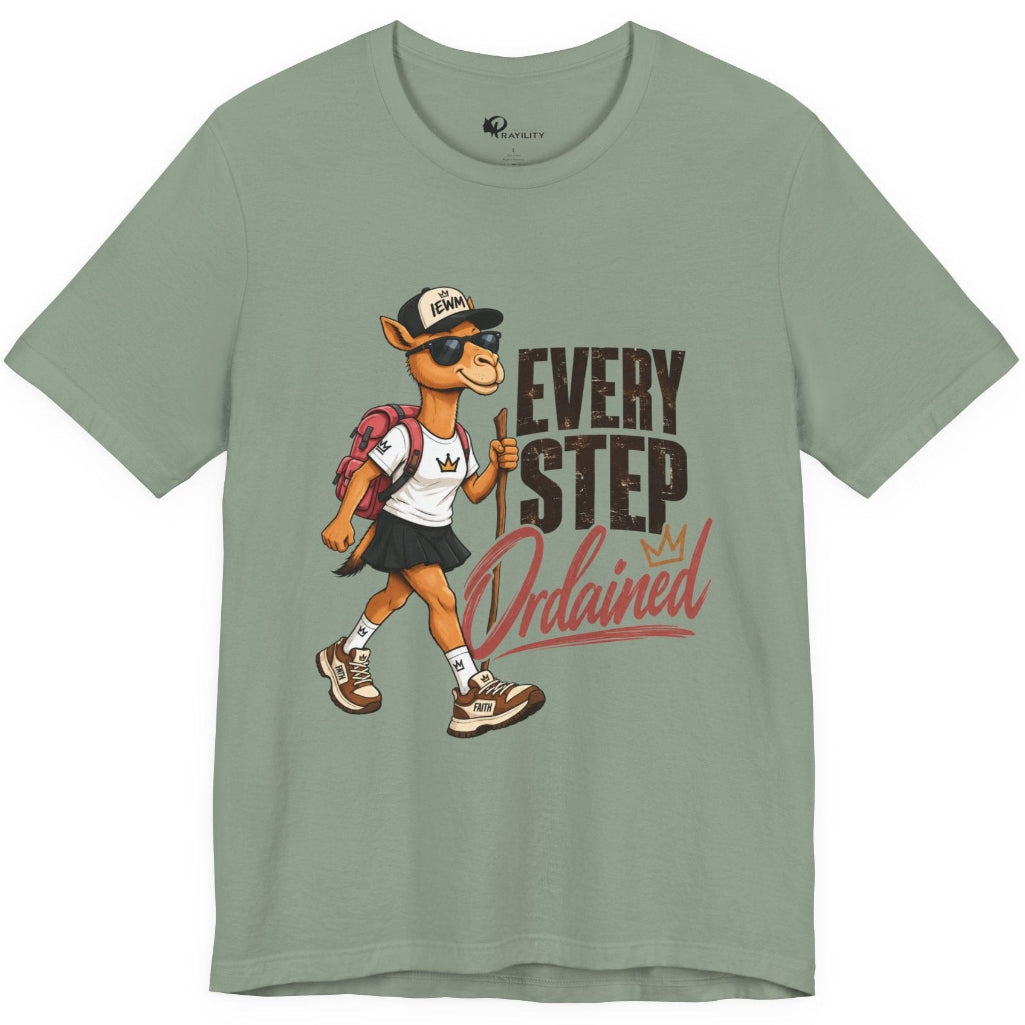 A sage green short-sleeve t-shirt with a graphic of a walking camel wearing a baseball cap and backpack, next to the text 'EVERY STEP Ordained'.