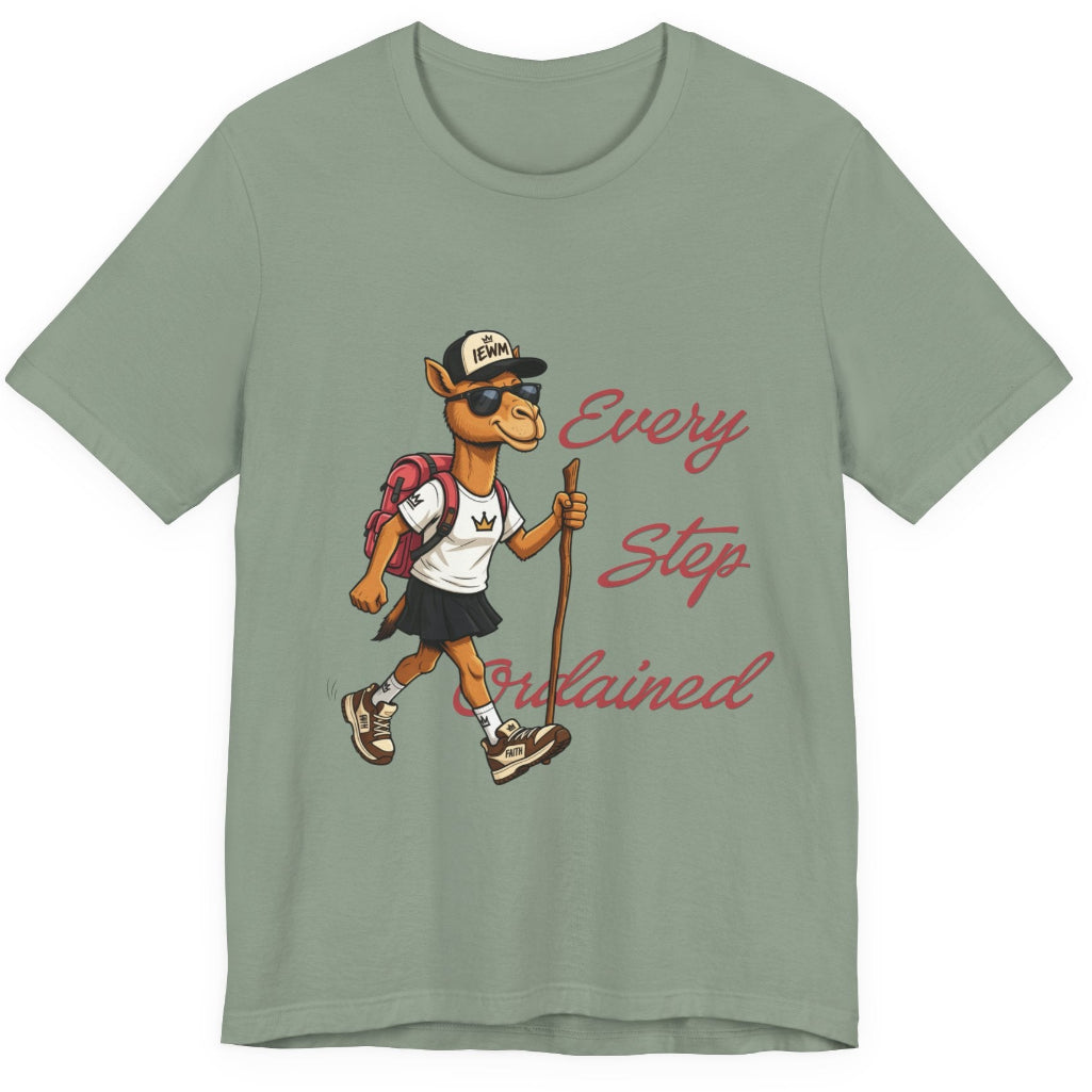 A sage green short-sleeve t-shirt with a graphic of a cartoon camel hiker wearing a white shirt, black skirt, and a backpack, next to the cursive text 'Every Step Ordained'.