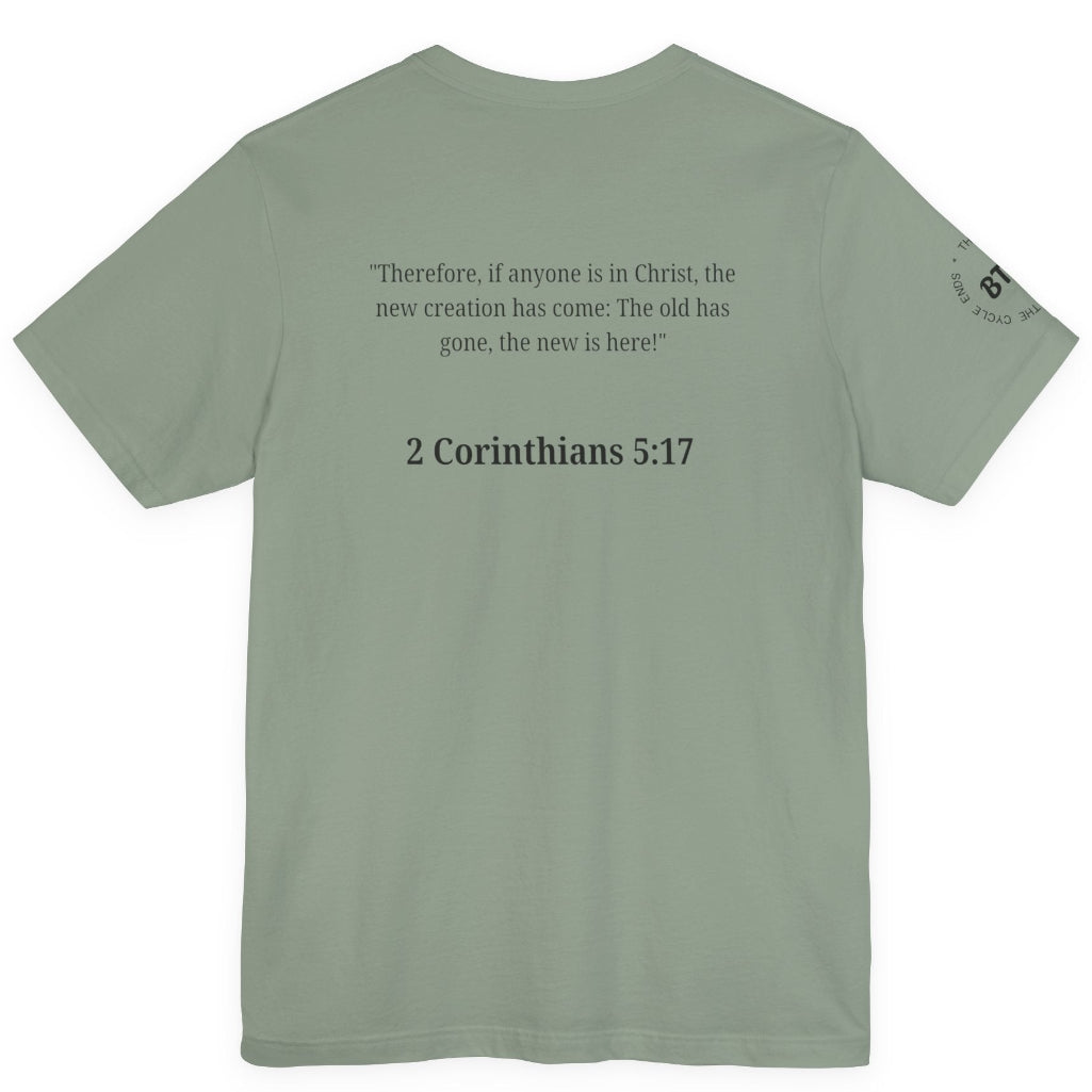 A sage green short-sleeve t-shirt shown from the back, featuring a black printed Bible verse from 2 Corinthians 5:17 and a small circular graphic on the left sleeve, displayed against a solid white background.