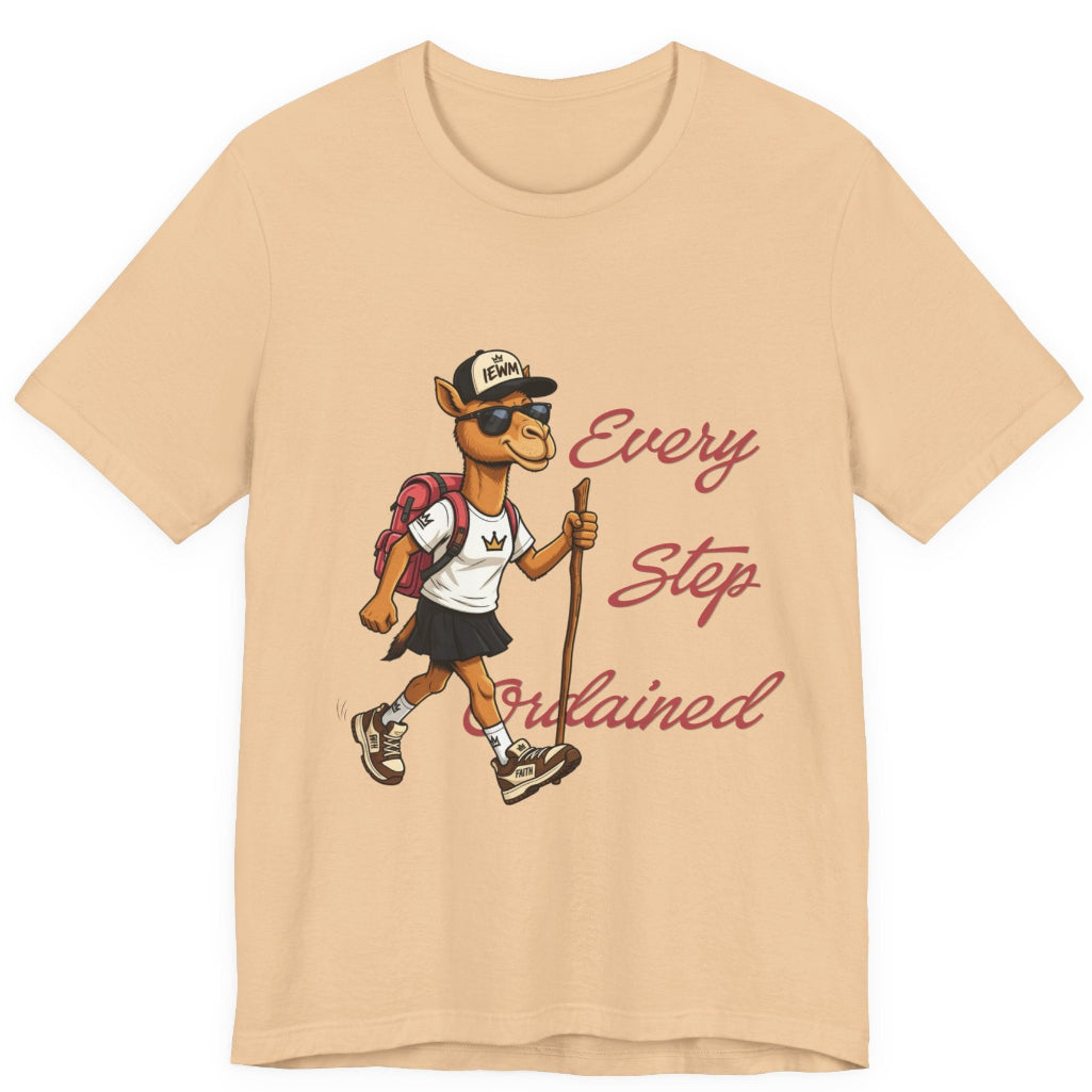 A sand colored short-sleeve t-shirt with a graphic of a cartoon camel hiker wearing a white shirt, black skirt, and a backpack, next to the cursive text 'Every Step Ordained'.
