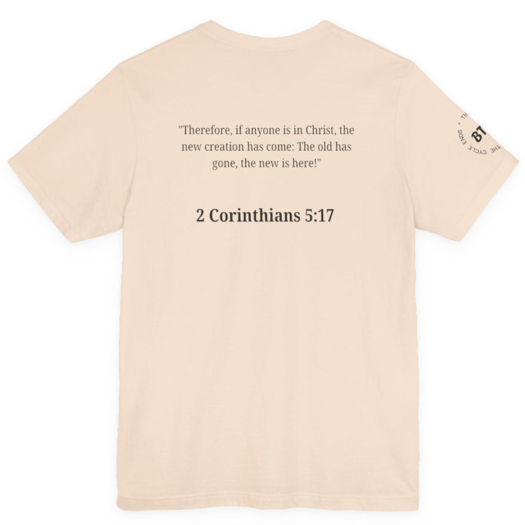 A light beige crewneck t-shirt shown from the back, featuring a black printed Bible verse from 2 Corinthians 5:17 in the center and a small circular graphic on the left sleeve.