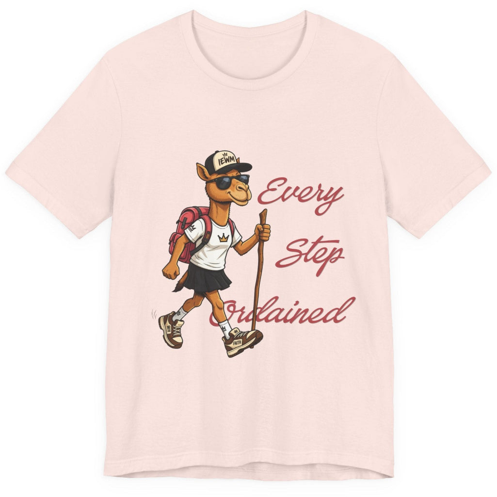 A soft pink short-sleeve t-shirt with a graphic of a cartoon camel hiker wearing a white shirt, black skirt, and a backpack, next to the cursive text 'Every Step Ordained'.
