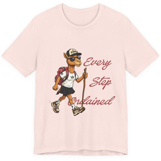A soft pink short-sleeve t-shirt with a graphic of a cartoon camel hiker wearing a white shirt, black skirt, and a backpack, next to the cursive text 'Every Step Ordained'.