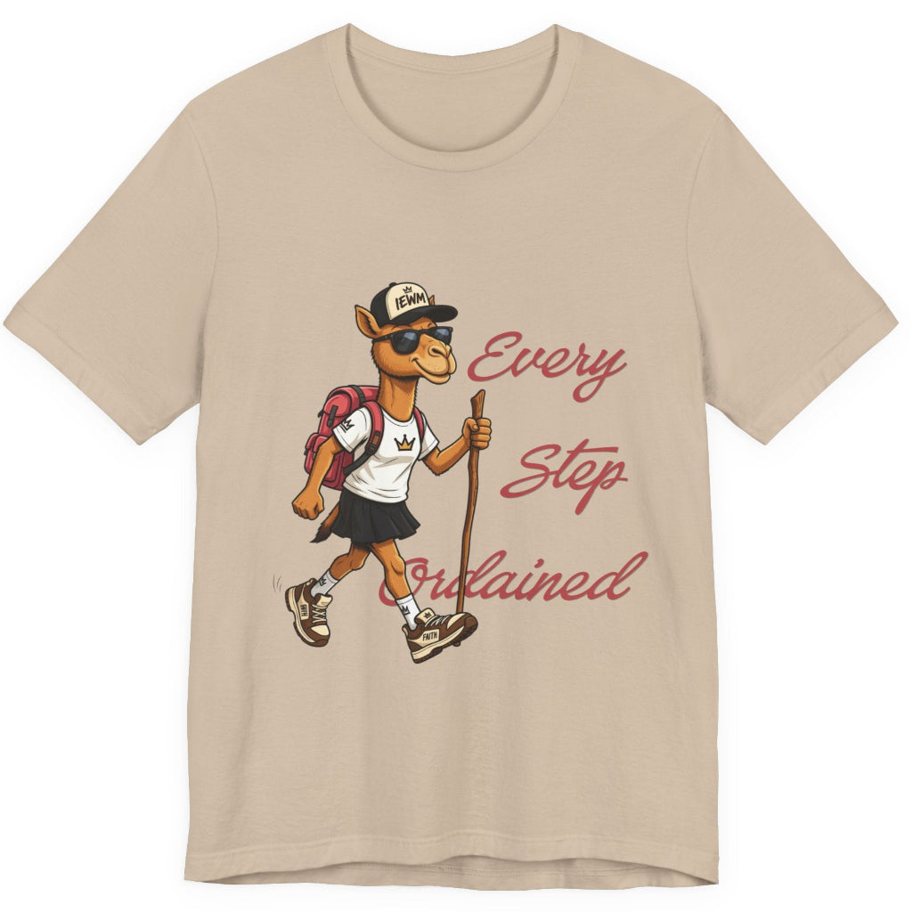 A tan short-sleeve t-shirt with a graphic of a cartoon camel hiker wearing a white shirt, black skirt, and a backpack, next to the cursive text 'Every Step Ordained'.