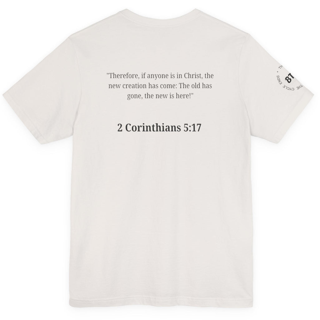 An off-white short-sleeve t-shirt shown from the back, featuring a black printed Bible verse from 2 Corinthians 5:17 in the center and a small circular graphic on the left sleeve.
