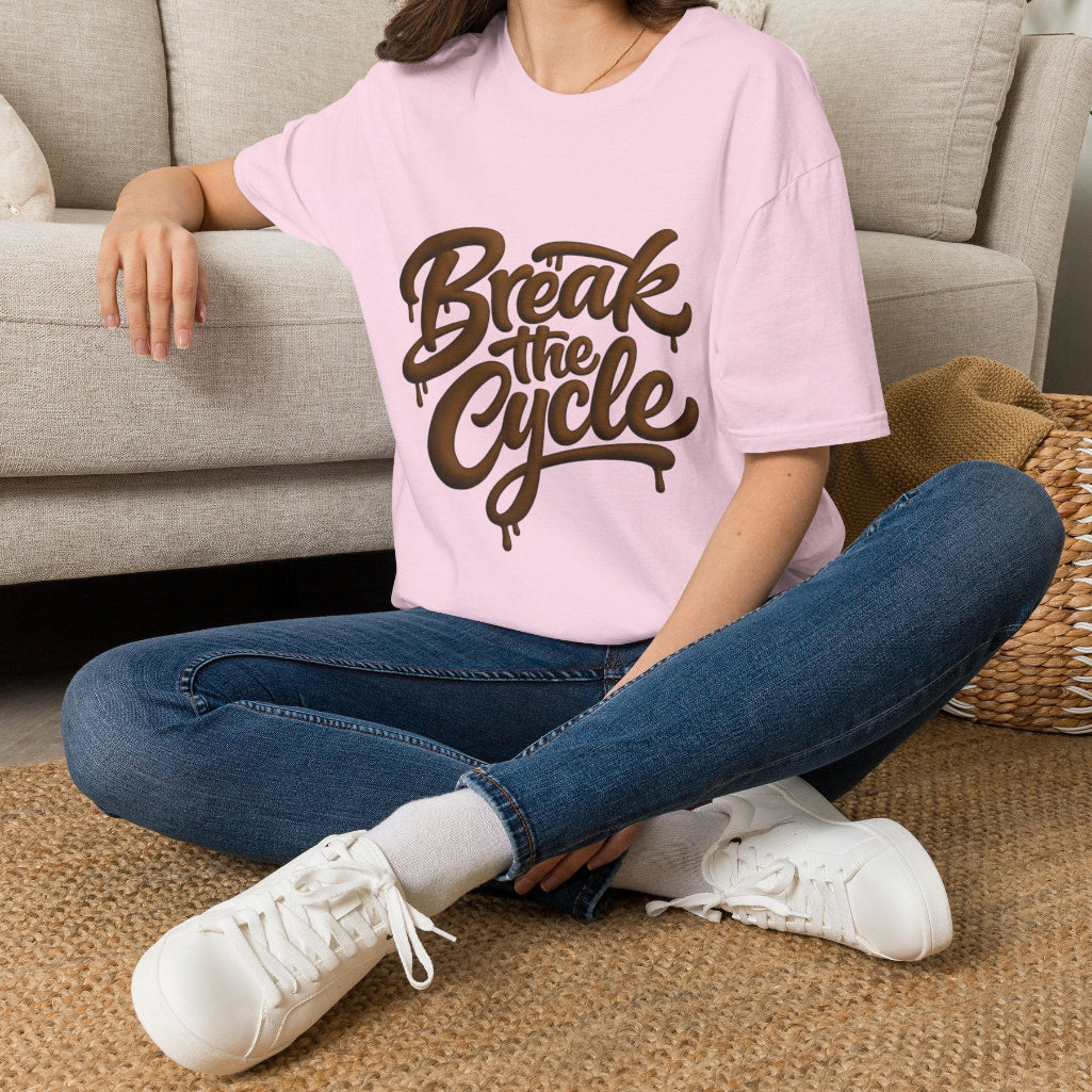 A woman sitting cross-legged on a woven rug in front of a light beige sofa, wearing a light pink oversized T-shirt with a brown 'Break the Cycle' graphic, blue jeans, and white sneakers.
